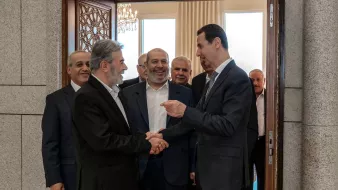 در این تصویر، چند مرد در یک اتاق ملاقات کرده‌اند. در جلو، دو نفر رو به روی هم ایستاده‌اند و با هم دست می‌دهند. فردی که سمت راست است ظاهراً در حال صحبت کردن و لبخند زدن است. دیگر افراد در پس‌زمینه نیز به این دو نفر نگاه می‌کنند و به نظر می‌رسد که در حال گفتگو یا مکالمه‌ای دوستانه هستند. فضا دارای نور طبیعی خوبی است و طراحی داخلی اتاق مدرن و شیک به نظر می‌رسد.