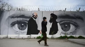 در این تصویر یک مرد در حال راه رفتن در کنار دیواری با یک نقاشی بزرگ است. دیوار دارای تصاویری از دو مرد است که به نظر می‌رسد در حال گفت‌وگو هستند. این نقاشی شامل چشمان بزرگی است که به وضوح بر روی دیوار کشیده شده و احساس عمیقی از توجه و نظارت را منتقل می‌کند. محیط اطراف خیابانی است و مرد به نظر می‌رسد مشغول صحبت کردن با تلفن همراهش باشد. طراحی و رنگ‌ها نسبتاً ملایم و معاصر به نظر می‌رسند.
