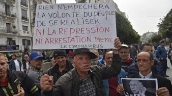 Sur l'image, un groupe de personnes se rassemble dans une rue, probablement lors d'une manifestation. Un homme tient une grande pancarte sur laquelle est écrit : "Rien n'empêchera la volonté du peuple de se réaliser ni la répression ni arrestation même le coronavirus". L'assemblée semble déterminée, et des expressions sérieuses sont visibles sur les visages des manifestants. L'ambiance évoque une lutte pour des droits ou des changements sociaux, sur fond de contestation.