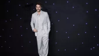 L'image montre un homme portant un costume léger à rayures, composé d'une veste et d'un pantalon ample. Il a une moustache et un sourire, et se tient debout avec une pose décontractée. L'arrière-plan est sombre, parsemé de petites lumières blanches qui ajoutent un effet élégant à la scène.