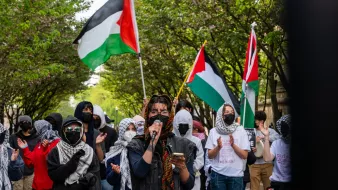 L'image montre une manifestation en plein air où plusieurs personnes sont rassemblées. Au premier plan, une personne prend la parole avec un microphone, vêtue de vêtements sombres et entourée de manifestants portant des masques. Beaucoup des présents arbore des foulards ou keffiehs. En arrière-plan, des drapeaux palestiniens flottent, ajoutant de la couleur à la scène. Les manifestants semblent soutenir une cause, probablement liée à la Palestine, et il y a une atmosphère de solidarité parmi le groupe. Les arbres en fleurs encadrent la scène, créant un contraste avec l'intensité de la manifestation.