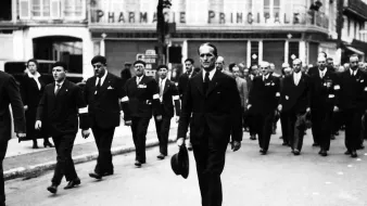 L'image montre un groupe de personnes défilant dans une rue, probablement lors d'un événement formel ou d'une commémoration. Au premier plan, un homme en costume noir porte un chapeau et marche avec assurance. Les autres participants portent aussi des costumes et certains semblent avoir des insignes ou des decorations. En arrière-plan, on peut voir un bâtiment avec l'enseigne "PHARMACIE PRINCIPALE". L'atmosphère générale de la scène semble sérieuse et respectueuse.