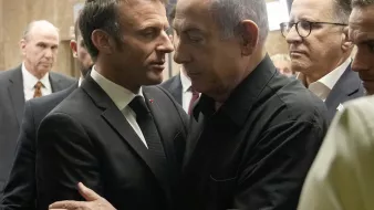 The image depicts two men in close proximity, engaged in a serious conversation. One man is dressed in a formal suit and tie, while the other is wearing a black shirt. They appear to be in an indoor setting, surrounded by several other individuals in the background, who are also dressed in formal attire. The atmosphere looks tense or important, suggesting a significant discussion is taking place.