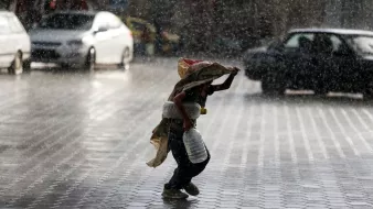 L'image montre un enfant courant sous la pluie intense, portant une écuelle en métal et un sac. Il semble se protéger de la pluie avec un tissu qui couvre sa tête et ses épaules. Le sol est mouillé et réfléchit la lumière, tandis que l'environnement autour de lui est flou en raison des cordes de pluie. Des voitures sont visibles en arrière-plan, accentuant l'atmosphère urbaine de la scène.