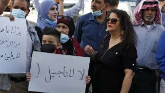 تظهر في الصورة مجموعة من الناس في مظاهرة. يرفع بعضهم لافتات، بينما يرتدي الآخرون كمامات. في المقدمة، يوجد طفل صغير يحمل لافتة مكتوب عليها "لا للتأجيل!"، وتحيط به مجموعة من البالغين، بينهم امرأة ترتدي فستانًا أسود وبعض الرجال بملابس تقليدية. تعكس الصورة جوًا من الاحتجاج والتعبير عن الرأي.