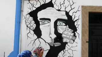 L'image montre une fresque murale en noir et blanc représentant un visage humain au milieu d'une apparente fissure dans le mur. Le visage a des traits marqués et semble exprimer une émotion intense. Des mains émergent des fissures, comme si elles tentaient de sortir. Au premier plan, deux femmes en hijab marchent, apportant une touche de couleur et de vie à la scène. Le tout crée un contraste entre l'art mural et la réalité environnante.
