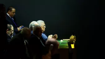 L'image montre un groupe d'hommes assis à une table, apparemment en train d'assister à un événement ou une réunion. L'éclairage est tamisé, mettant en évidence certaines personnes tandis que d'autres sont dans l'ombre. L'un des hommes est occupé avec son téléphone portable. Les visages semblent préoccupés et concentrés, donnant une impression de gravité et d'importance à la situation.