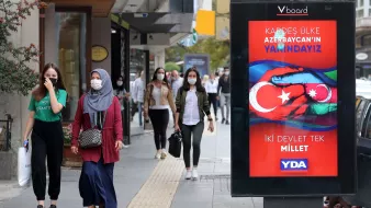 L'image montre une scène de rue animée où des personnes marchent sur un trottoir. À gauche, deux femmes avancent, l'une portant un hijab gris et l'autre en tenue décontractée. À droite, un panneau d'affichage numérique affiche un message en turc, avec des couleurs vives et des symboles représentant les drapeaux de la Turquie et de l'Azerbaïdjan. Le texte semble faire référence à un soutien entre les deux pays. Plusieurs passants peuvent être aperçus en arrière-plan, portant tous des masques de protection.