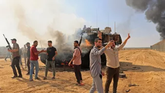 تظهر الصورة مجموعة من الأشخاص في منطقة صحراوية تتصاعد فيها الدخان من عربة مدرعة مدمَّرة. بعض الأشخاص يرفعون أيديهم في إشارة النصر أو الاحتفاء، بينما يبدو آخرون مشغولين بالتفاعل مع بعضهم البعض. الأجواء تعكس حالة من التوتر، مع وجود عمود من الدخان الأسود في الخلفية. السماء زرقاء ولكن مشوشة بسبب الدخان.