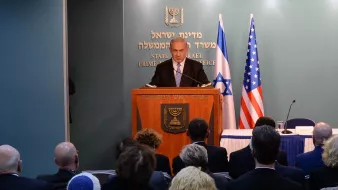 L'image montre un homme en train de s'adresser à un public depuis un podium marqué du sceau d'Israël et des États-Unis. Il y a des personnes assises derrière lui, écoutant son discours. On peut voir des drapeaux israélien et américain en arrière-plan. L'ambiance semble formelle, probablement dans le cadre d'une conférence ou d'une réunion officielle.