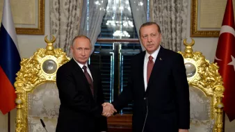 L'image montre deux hommes se serrant la main dans un cadre formel. Ils sont probablement en train de discuter d'accords ou de collaborations. L'un d'eux semble être en costume sombre avec une cravate, tandis que l'autre porte également un costume et une cravate. Ils se tiennent devant des fauteuils élégants et ornés, avec des drapeaux en arrière-plan, ce qui suggère un contexte officiel ou diplomatique. La lumière naturelle entre par les fenêtres, créant une atmosphère importante.
