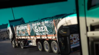 L'immagine mostra un camion lungo che trasporta una grande quantità di beni. Il veicolo è visibile da un angolo diverso, suggerendo che la foto potrebbe essere stata scattata da un altro veicolo. Sulla superficie del camion è presente un'informazione, probabilmente un messaggio di solidarietà o supporto, che menziona "Gaza" e "marzo 2024". Il contesto suggerisce un'operazione umanitaria o di aiuto. Sullo sfondo si possono vedere elementi di un paesaggio stradale.