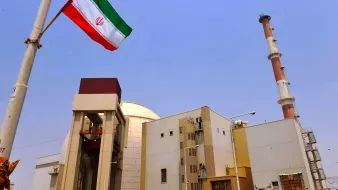 L'image montre une installation industrielle avec des structures modernes. On peut voir une grande cheminée au fond et un bâtiment principal qui semble être lié à l'énergie. Au premier plan, il y a un mât avec le drapeau iranien flottant, qui arbore des bandes rouges, blanches et vertes. Le ciel est dégagé, créant un contraste avec les bâtiments. L'ensemble évoque une infrastructure liée à la production d'énergie, probablement nucléaire.