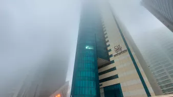 L'image montre un paysage urbain avec des gratte-ciels partiellement cachés par un épais brouillard. La visibilité est réduite, ce qui donne une atmosphère mystérieuse et peu lumineuse. Les bâtiments ont des façades modernes, avec des reflets de verre. On peut distinguer des éléments architecturaux, mais l'intensité du brouillard rend difficile l'observation claire des détails. Les lumières des rues sont visibles, créant un contraste avec l'environnement brumeux.