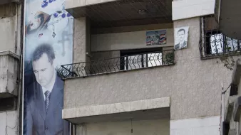 L'immagine mostra un edificio con muri decorati da grandi striscioni e manifesti. C'è un grande ritratto di una figura maschile su un muro, mentre altri manifesti e striscioni sono esposti sulla facciata dell'edificio e sul balcone. Alcuni uomini si trovano sopra al balcone, visibili mentre osservano l'esterno. Il contesto suggerisce un ambiente urbano con elementi di sostegno e celebrazione, magari legati a un leader o a eventi politici.