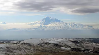 L'image montre un paysage montagneux impressionnant, avec un grand sommet enneigé qui semble être un volcan. Le sommet est entouré de nuages légers, créant une atmosphère mystique. Au premier plan, des collines aux teintes terreuses et parfois enneigées s'étendent, ajoutant à la beauté naturelle de la scène. Le ciel est partiellement couvert, laissant transparaître une lumière douce qui éclaire le paysage.