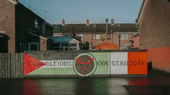 L'image montre un mur peint avec une fresque qui illustre un message de solidarité ou de lutte. On peut y voir des éléments graphiques comme des drapeaux, symbolisant probablement des nations ou des groupes. Les couleurs rouge, vert et blanc sont présentes, évoquant des thèmes de culture ou d'appartenance. Le sol est mouillé, reflétant les bâtiments environnants et créant une atmosphère calme malgré le message fort du graffiti.