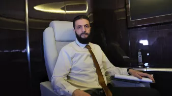 الصورة تُظهر شخصًا جالسًا في مقعد مريح داخل مكان يبدو راقيًا. يرتدي قميصًا فاتحًا ورابطة عنق، ويبدو في وضعية تفكر. خلفه توجد شاشة ومكتب أنيق، مما يوحي بأجواء مهنية.