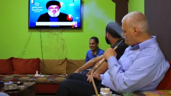 L'image montre un groupe d'hommes assis dans un salon avec des murs verts. Ils semblent regarder la télévision, où l'on peut voir un personnage à l'écran. L'un des hommes fume une pipe à eau, tandis que les autres sont engagés dans la conversation ou en observant l'écran. L'environnement est décontracté, avec des canapés confortables et des éléments de décoration simples.