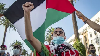 L'image montre une manifestation où un homme brandit fièrement un drapeau palestinien. Il porte un masque et est entouré de palmiers, ce qui indique un environnement ensoleillé. Son expression montre une détermination forte, et il semble engagé dans une cause importante. D'autres personnes sont présentes dans le fond, ajoutant au contexte collectif de la protestation.