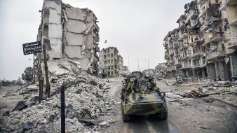 L'image montre une scène de destruction urbaine, probablement à la suite d'un conflit. On peut voir des bâtiments en ruine, avec des façades effondrées et des débris éparpillés sur le sol. Au premier plan, il y a un véhicule militaire, sur lequel des soldats sont présents. L'atmosphère semble sombre et chargée, soulignant les ravages causés par la guerre.