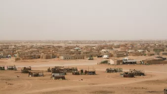 L'image montre un paysage désertique, probablement dans une région aride. On peut voir une vaste étendue de terre sèche, avec des habitations simples construites en terre et en matériaux naturels, disséminées dans le paysage. Certaines constructions semblent être regroupées, tandis que d'autres sont isolées. L'atmosphère est probablement chaude et poussiéreuse, avec un ciel clair ou légèrement embrumé. Les couleurs dominantes sont des tons de beige, de marron et de jaune, typiques des déserts.