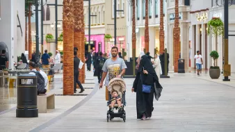 L'image montre une scène animée dans un centre commercial. On peut voir des palmés en arrière-plan et des magasins de différentes boutiques. Un homme marche avec un enfant dans une poussette, tandis qu'une femme, vêtue d'une abaya, les accompagne. Plusieurs autres personnes sont visibles dans l'allée, créant une ambiance de vie urbaine dynamique. La lumière est claire et l'atmosphère semble accueillante.