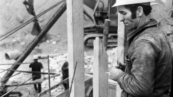 L'image montre un homme en train de travailler sur un chantier de construction. Il porte un casque de protection blanc et semble concentré sur sa tâche. À proximité, on peut voir des éléments en béton vertical, probablement des murs ou des supports. En arrière-plan, des ouvriers sont également visibles, et une machine lourde est à l'œuvre. L'image est en noir et blanc, ce qui lui donne un aspect rétro et saisissant. L'ambiance générale évoque le travail manuel et l'effort dans le domaine de la construction.