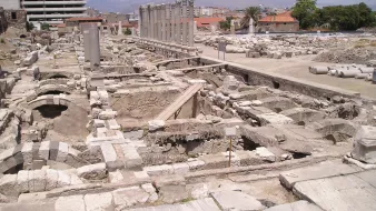 L'image montre un site archéologique avec des ruines et des structures anciennes. On aperçoit des colonnes dressées, probablement d'un bâtiment antique, ainsi que des fondations de murs en pierre. En arrière-plan, on peut distinguer des bâtiments modernes, contrastant avec les vestiges historiques. Le ciel est clair et le paysage est légèrement vallonné au loin. L'endroit semble être une zone de fouilles, avec des éléments qui témoignent d'une ancienne civilisation.