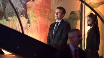 L'image montre un homme en costume, debout sur une scène, avec une femme à ses côtés qui porte un microphone et un ouïe. L'arrière-plan présente des couleurs et des formes abstraites, suggérant un environnement artistique ou un événement important. L'homme semble attentif, tandis que la femme semble concentrée sur la situation. Il y a également un autre homme en arrière-plan qui marche, suggérant une ambiance dynamique.