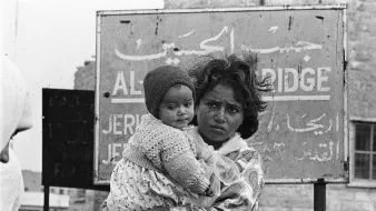 این تصویر یک کودک را نشان می‌دهد که در آغوش یک زن ایستاده است. زن ظاهراً نگران و جدی به نظر می‌رسد و کودک در آغوشش پایش را به دور او حلقه کرده است. پس‌زمینه تصویر شامل یک تابلو با نوشته‌ها به زبان عربی و انگلیسی است که نام مکانی را نشان می‌دهد. حالت چهره و لباس‌های آن‌ها نشان‌دهنده سختی‌های احتمالی در زندگی است. تصویر به وضوح وضعیت انسانی و احساسات عمیق را منتقل می‌کند.