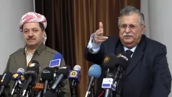 L'image montre deux hommes en conférence de presse, entourés de plusieurs micros et caméras provenant de divers médias. L'homme à gauche porte une tenue traditionnelle kurde avec un foulard, tandis que l'homme à droite, habillé en costume, semble s'adresser à l'audience en pointant du doigt. L'ambiance est formelle, indiquant un moment important de communication ou d'annonce.