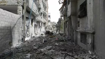 L'image montre une rue dévastée, probablement dans une zone de conflit. Les bâtiments de chaque côté sont endommagés, avec des murs effondrés et des débris éparpillés sur le sol. Des fils électriques pendent, et l'atmosphère générale dégage une impression de destruction et d'abandon. On peut observer des traces des conséquences des affrontements, avec un paysage urbain sévèrement altéré.