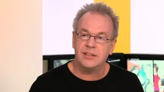 L'image montre un homme avec des cheveux gris et des lunettes. Il porte un t-shirt noir et semble en train de parler, avec un fond flou qui suggère un environnement de discussion ou d'interview.
