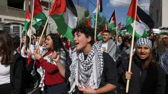 L'image montre une foule de manifestants, principalement des jeunes, participant à une marche ou une manifestation. Ils portent des drapeaux palestiniens aux couleurs rouge, noir, blanc et vert, et certains portent des keffiehs, des écharpes traditionnelles palestiniennes, souvent à motifs noirs et blancs. Les manifestants semblent passionnés et chantent, exprimant leur soutien à une cause. En arrière-plan, on aperçoit des bâtiments et un ciel partiellement nuageux.