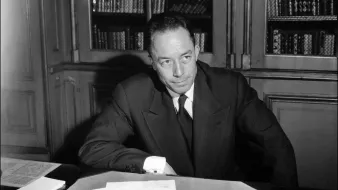 L'image montre un homme assis à un bureau, avec des documents éparpillés devant lui. Il est habillé de manière formelle, portant un costume sombre et une cravate. L'arrière-plan présente des étagères remplies de livres. L'éclairage met en valeur ses traits, suggérant un moment de réflexion ou de concentration.