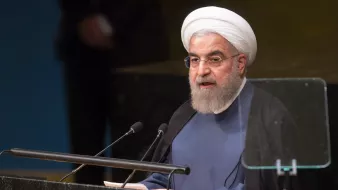 The image depicts a man delivering a speech at a podium. He has a distinctive white turban and a beard, dressed in a dark robe. The setting appears to be a formal event, likely at a conference or assembly, given the backdrop and the podium's design. The focus is on him as he addresses an audience, emphasizing the seriousness and importance of the moment.