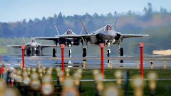 The image features three F-35 fighter jets on an airstrip, showcasing their sleek and modern designs. The jets are positioned closely together, and the background features a blurred green landscape, indicating a rural setting. The foreground includes several lights lining the runway, which adds a sense of depth to the scene. The overall atmosphere appears dynamic and focused on military aviation.
