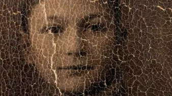 L'image représente un portrait d'une personne, probablement un jeune garçon, dont les traits sont légèrement obscurcis par un certain vieillissement et des fissures à la surface. Le visage montre des expressions délicates et le regard engageant, mais les détails sont atténués à cause de l'état usé de la photographie. Le fond semble sombre, ce qui accentue l'apparence vieillie de l'image.