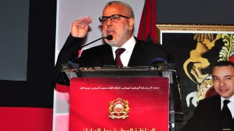 L'image montre un homme en train de s'exprimer lors d'un événement officiel. Il porte des lunettes et une veste noire, et il semble faire un geste de salutation avec sa main. Derrière lui, un panneau avec du texte en arabe et français est visible, ainsi qu'une photo d'un autre homme, probablement une figure politique. Le contexte semble être lié à des discussions sur des questions fiscales.