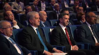L'image montre un groupe d'hommes assis dans une salle de conférence, apparemment lors d'un événement officiel. Ils portent des costumes sombres, avec une attention particulière sur deux hommes au premier plan, l'un en costume noir avec une cravate bleue, et l'autre en costume noir avec une cravate rouge. En arrière-plan, on peut voir d'autres personnes, probablement des participants ou des invités, attentifs à ce qui se passe sur scène. L'atmosphère semble formelle et sérieuse.