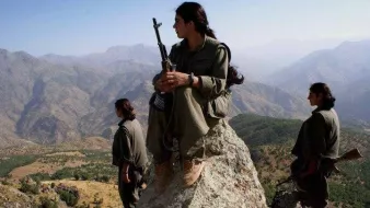 L'image montre trois personnes, probablement des combattants, dans un paysage montagneux. L'une d'elles est assise sur une pierre, tenant une arme, avec les jambes repliées. Les deux autres se tiennent debout en arrière-plan, également armées. Le décor est naturel, avec des montagnes majestueuses et une plaine en dessous, suggérant un environnement rural et sauvage. Les combattants portent des vêtements pratiques adaptés à leur environnement. L'ambiance de la scène semble sérieuse et déterminée.