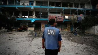 L'image montre un homme portant un gilet de l'ONU, observant un bâtiment endommagé. Le bâtiment a des murs peints et des balcons, mais il est en ruine, avec des fenêtres brisées et des débris éparpillés autour. L'atmosphère est sombre, suggérant une situation de conflit ou de crise, avec d'autres personnes visibles en arrière-plan dans un environnement chaotique.
