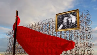 L'image représente une scène créative et symbolique. On y voit une structure composée de fils en métal enroulés, formant un mur. Devant ce mur, un tissu rouge flotte, suggérant une dynamique de mouvement et de vie. Accrochée au mur, une image en noir et blanc dans un cadre doré attire l'attention, ajoutant une dimension artistique à la composition. Le ciel en arrière-plan présente des nuages épars, offrant un contraste entre la rigidité de la structure et la légèreté du tissu. L'ensemble dégage une ambiance à la fois poétique et contemplative.