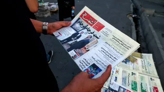 L'image montre une personne tenant un journal ouvert. Le titre est bien visible et semble relater des informations d'actualité, potentiellement politiques ou sociales. En arrière-plan, on aperçoit un kiosque à journaux avec d'autres exemplaires de journaux exposés. L'environnement semble urbain, avec des voitures garées et un sol en pavé.