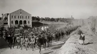 L'image montre une troupe de soldats à cheval marchant en formation le long d'un chemin. En arrière-plan, on peut voir des bâtiments en pierre, dont une grande structure avec des fenêtres. Le sol est poussiéreux, et on remarque également quelques personnes à pied, dont une femme en robe. L'atmosphère semble être celle d'une époque historique, probablement d'un conflit ou d'une quelconque mobilisation militaire. Les soldats portent des uniformes similaires et semblent déterminés dans leur avancée.