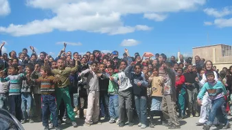 تظهر في الصورة مجموعة كبيرة من الأطفال والشباب يتجمعون في مكان واحد، يبدو أنهم متحمسون ويقومون بالتفاعل مع شيء ما، ربما يجتمعون لمتابعة حدث أو نشاط معين. السماء في الخلفية زرقاء مع بعض السحب، والمكان يبدو كأنه في منطقة حضرية. يرتدي الأطفال ملابس متنوعة، وتعكس تعبيراتهم الحماس والإثارة.