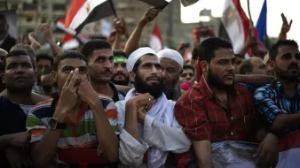 L'image montre un groupe de personnes rassemblées lors d'une manifestation. On peut voir des hommes de différentes origines, certains portant des vêtements traditionnels, d'autres des t-shirts colorés. Ils semblent tous attentifs et engagés, certains levant les mains ou tenant des drapeaux. L'ambiance semble être celle d'une mobilisation ou d'une expression de soutien pour une cause. Le fond montre un environnement urbain, avec d'autres manifestants et des drapeaux visibles.
