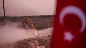L'image montre un tank, probablement militaire, se déplaçant sur une route poussiéreuse. Au premier plan, on aperçoit un drapeau turc, qui ajoute un contexte national à la scène. L'arrière-plan présente un paysage rural, avec des champs, et l'on peut voir de la poussière se lever autour du tank, suggérant une atmosphère de mouvement ou d'activité militaire. L'éclairage est doux, créant une ambiance de crépuscule.