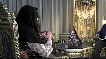 الصورة تظهر مشهداً بين شخصين يجلسان على كراسي مزخرفة. الشخص في الجهة اليسرى يرتدي ملابس تقليدية ويحمل بيديه بعض الأشياء. بينما الشخص في الجهة اليمنى يبدو أنه يقرا من كتاب أو ملاحظات. الخلفية تحتوي على زينة تقليدية، مع إضاءة ناعمة تعكس أجواء الحوار.